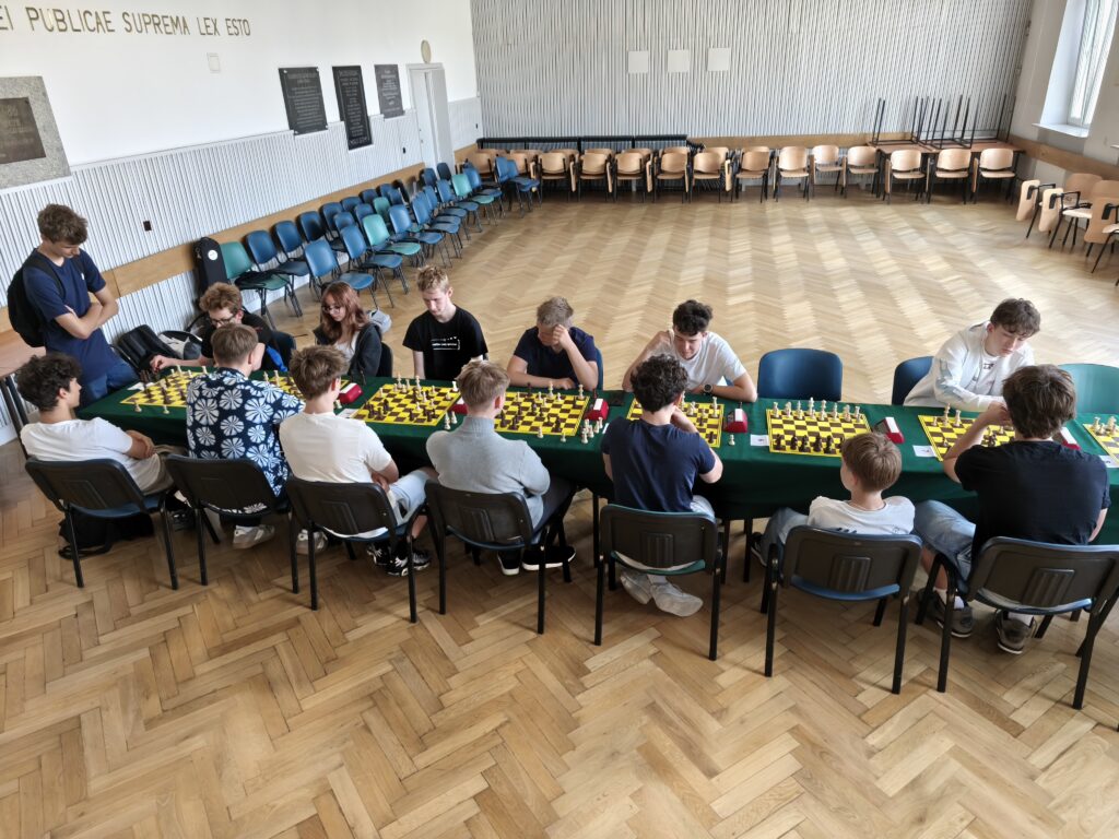 Szerokie ujęcie sali, w której odbywa się turniej szachowy. Kilku uczestników gra przy ustawionych stołach, a wokół sali ustawiono krzesła dla zawodników i obserwatorów. Fotografia pokazuje ogólną atmosferę i organizację wydarzenia.
