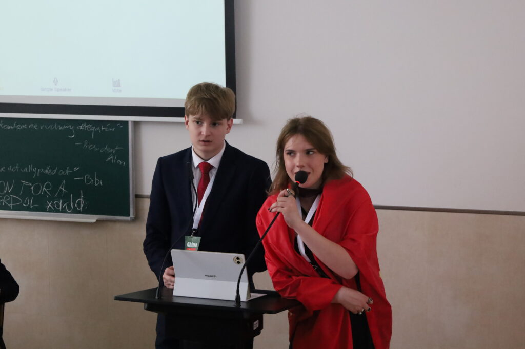 Dwoje uczestników prowadzi wystąpienie przy mównicy. Jedna osoba przemawia do mikrofonu, druga obsługuje tablet i materiały prezentacyjne, uczestnicząc w oficjalnej części debaty.