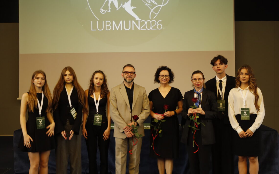 Grupa organizatorów, uczestników i opiekunów pozuje do wspólnego zdjęcia podczas konferencji LUBMUN 2026. Na ekranie za nimi widoczne jest logo wydarzenia, a część osób trzyma czerwone róże jako symbol podziękowania lub wyróżnienia.