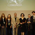 Grupa organizatorów, uczestników i opiekunów pozuje do wspólnego zdjęcia podczas konferencji LUBMUN 2026. Na ekranie za nimi widoczne jest logo wydarzenia, a część osób trzyma czerwone róże jako symbol podziękowania lub wyróżnienia.