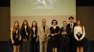 Grupa organizatorów, uczestników i opiekunów pozuje do wspólnego zdjęcia podczas konferencji LUBMUN 2026. Na ekranie za nimi widoczne jest logo wydarzenia, a część osób trzyma czerwone róże jako symbol podziękowania lub wyróżnienia.