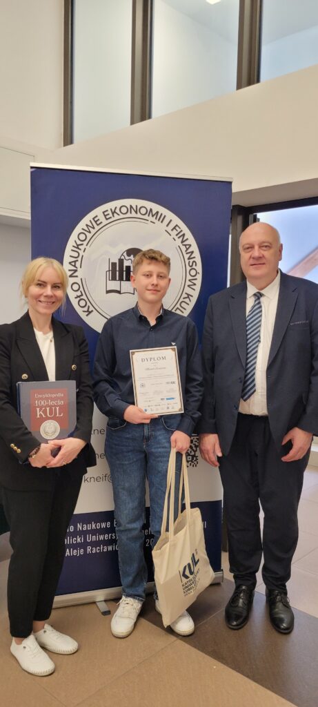 Laureat konkursu pozuje wspólnie z dwojgiem przedstawicieli uczelni. Trzyma dyplom i torbę KUL, a obok niego stoją kobieta z książką „Encyklopedia 100-lecia KUL” oraz mężczyzna w garniturze. W tle widoczny jest baner Koła Naukowego Ekonomii i Finansów.