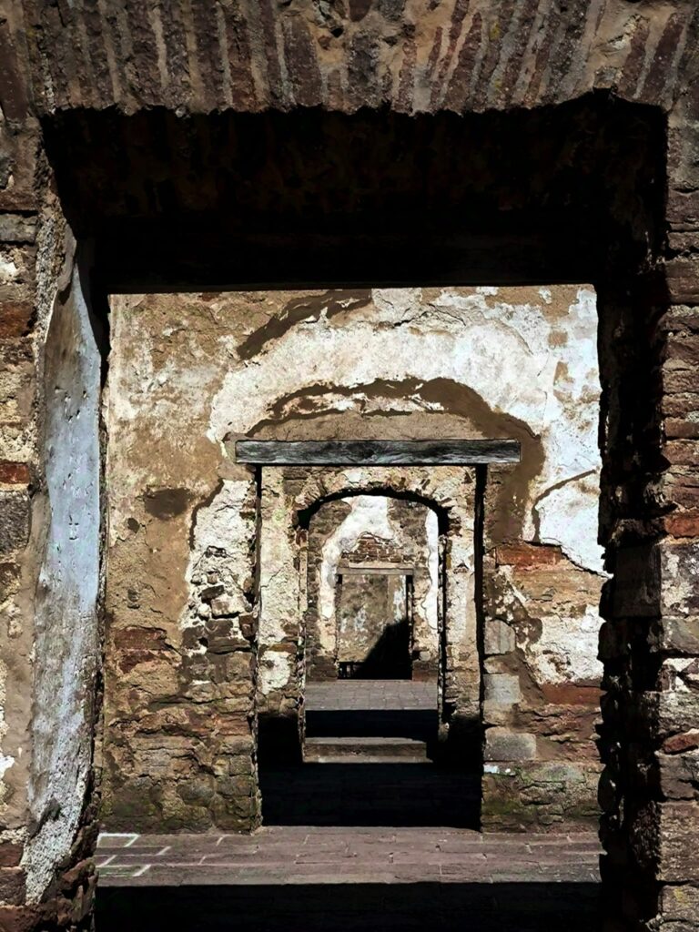 Widok przez serię kolejnych kamiennych przejść w ruinach historycznej budowli. Otwory drzwiowe ustawione są jeden za drugim, tworząc efekt głębokiej perspektywy. Ściany są częściowo zniszczone, z odsłoniętym kamieniem i cegłą.