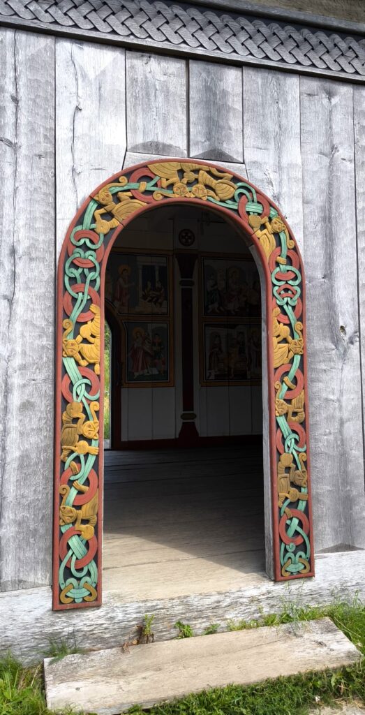 Ozdobione wejście do drewnianego budynku. Łukowata futryna pokryta jest kolorowymi ornamentami splecionych węzłów i stylizowanych zwierząt, nawiązującymi do sztuki nordyckiej lub wikińskiej. W głębi widoczne są malowane dekoracje wnętrza.