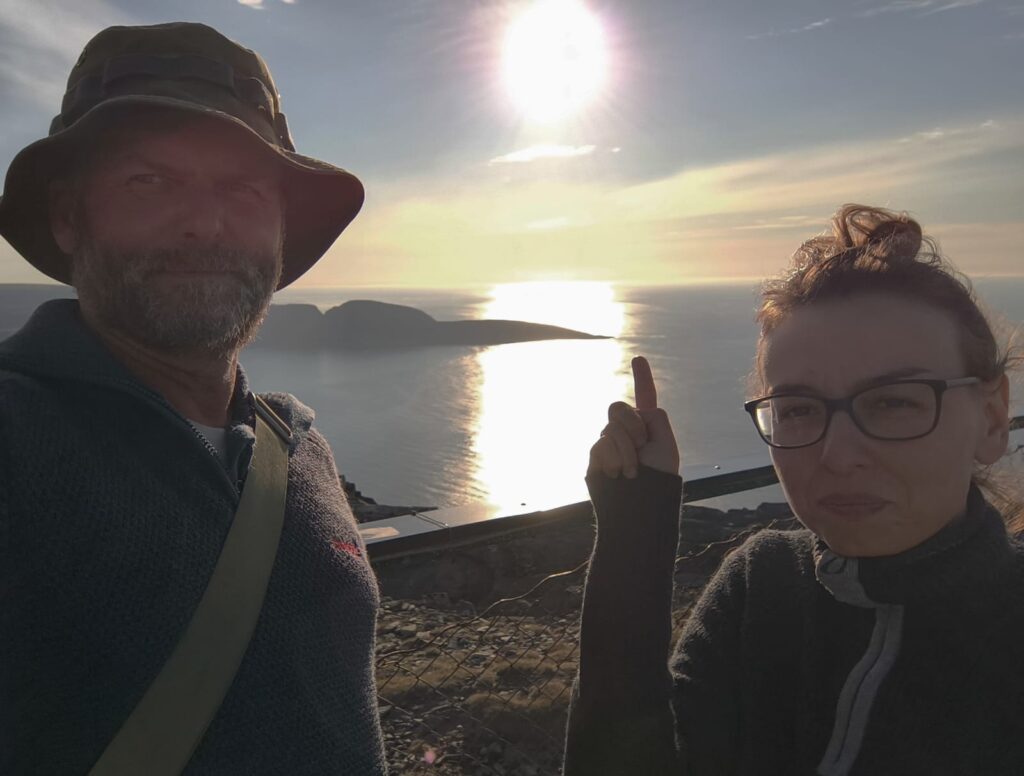Selfie dwóch osób wykonane na punkcie widokowym nad morzem. W tle nisko nad horyzontem świeci słońce, którego odbicie tworzy jasną smugę na wodzie. Jedna z osób wskazuje palcem w stronę zachodzącego lub nisko położonego słońca.