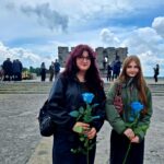 Dwie młode dziewczyny stoją na placu przed pomnikiem lub miejscem pamięci, trzymając w dłoniach niebieskie róże. W tle widoczne są schody, grupa uczestników uroczystości oraz fragment kamiennego monumentu. Nad całą sceną rozciąga się dramatyczne, zachmurzone niebo. Fotografia ma uroczysty i refleksyjny charakter, sugerując udział w wydarzeniu upamiętniającym lub rocznicowym.