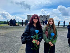 Dwie młode dziewczyny stoją na placu przed pomnikiem lub miejscem pamięci, trzymając w dłoniach niebieskie róże. W tle widoczne są schody, grupa uczestników uroczystości oraz fragment kamiennego monumentu. Nad całą sceną rozciąga się dramatyczne, zachmurzone niebo. Fotografia ma uroczysty i refleksyjny charakter, sugerując udział w wydarzeniu upamiętniającym lub rocznicowym.