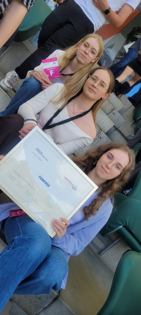 Trzy finalistki siedzą na trybunach. Jedna z nich trzyma duży certyfikat projektu „Fonfree”. Wszystkie mają identyfikatory wydarzenia i patrzą w stronę aparatu. Zdjęcie wykonano w jasnym, stadionowym otoczeniu.