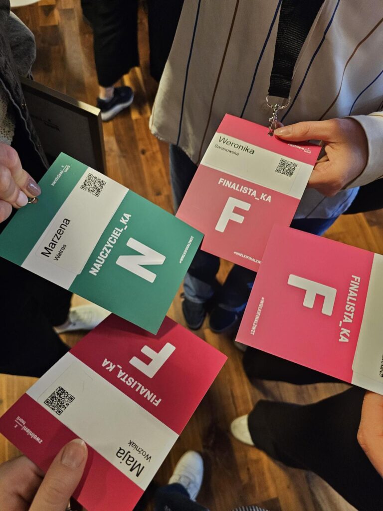 Uczestnicy wydarzenia stoją w kręgu i prezentują identyfikatory. Widoczne są różowe identyfikatory z napisem „FINALISTA_KA” oraz zielony identyfikator nauczycielki. Na identyfikatorach znajdują się imiona, kody QR i duże litery oznaczające role uczestników. Zdjęcie wykonano z góry, na tle drewnianej podłogi.
