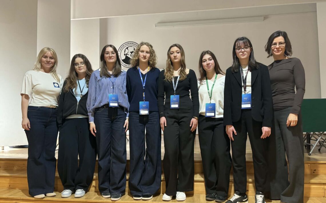 Grupowe zdjęcie wykonane na szkolnej scenie lub w auli. W jednym rzędzie stoi osiem kobiet – sześć uczennic lub uczestniczek projektu oraz dwie dorosłe opiekunki. Większość osób ma na szyi identyfikatory konferencyjne lub projektowe. Uczestniczki są ubrane w stonowane, głównie granatowe, czarne i białe stroje. W tle widoczna jest jasna ściana z logo szkoły, a całość ma charakter pamiątkowej fotografii wykonanej po prezentacji, konkursie lub wydarzeniu edukacyjnym.