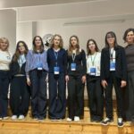 Grupowe zdjęcie wykonane na szkolnej scenie lub w auli. W jednym rzędzie stoi osiem kobiet – sześć uczennic lub uczestniczek projektu oraz dwie dorosłe opiekunki. Większość osób ma na szyi identyfikatory konferencyjne lub projektowe. Uczestniczki są ubrane w stonowane, głównie granatowe, czarne i białe stroje. W tle widoczna jest jasna ściana z logo szkoły, a całość ma charakter pamiątkowej fotografii wykonanej po prezentacji, konkursie lub wydarzeniu edukacyjnym.