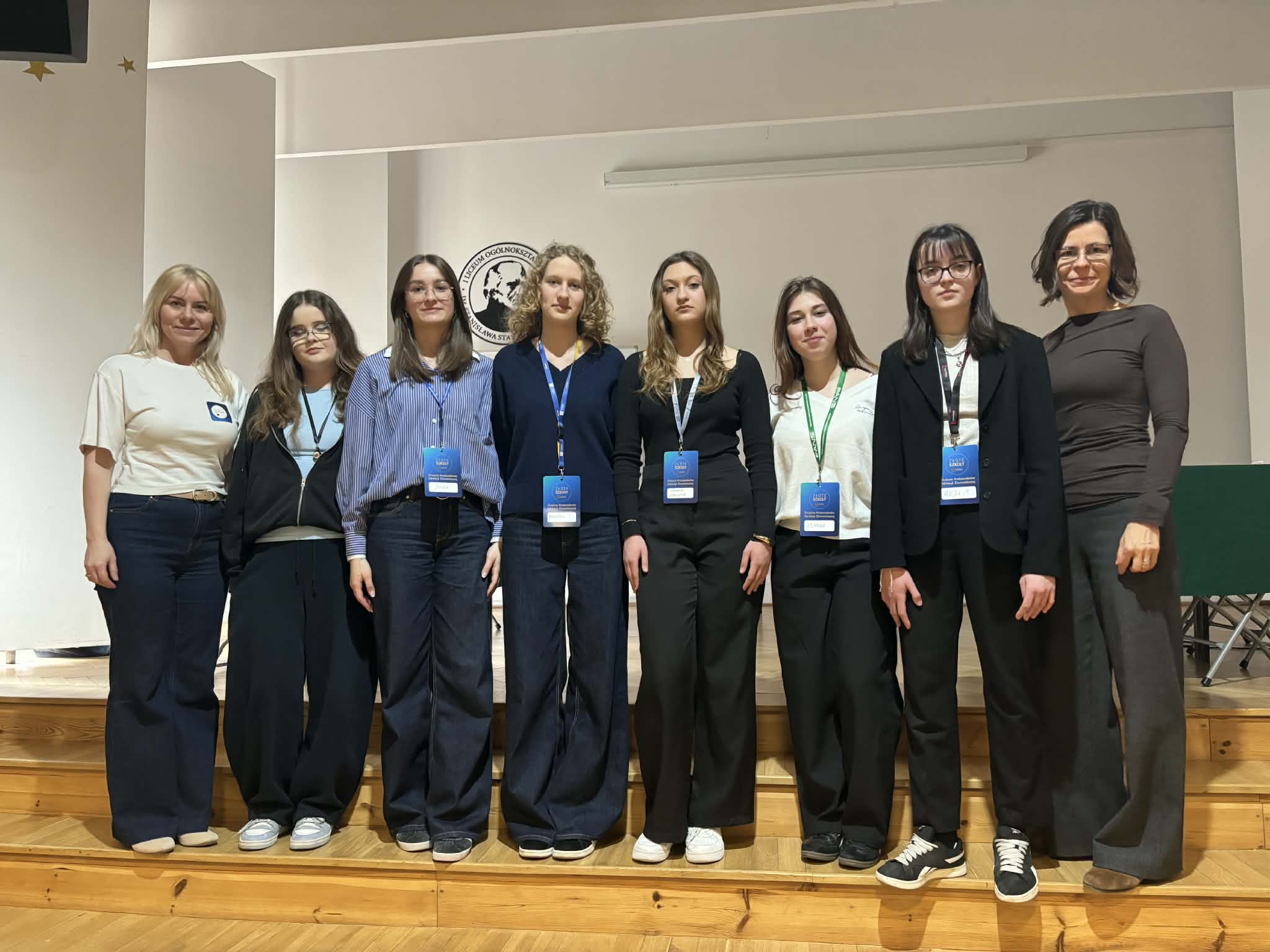 Grupowe zdjęcie wykonane na szkolnej scenie lub w auli. W jednym rzędzie stoi osiem kobiet – sześć uczennic lub uczestniczek projektu oraz dwie dorosłe opiekunki. Większość osób ma na szyi identyfikatory konferencyjne lub projektowe. Uczestniczki są ubrane w stonowane, głównie granatowe, czarne i białe stroje. W tle widoczna jest jasna ściana z logo szkoły, a całość ma charakter pamiątkowej fotografii wykonanej po prezentacji, konkursie lub wydarzeniu edukacyjnym.
