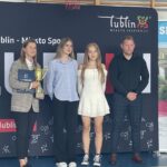 Cztery osoby pozują do zdjęcia podczas uroczystości sportowej w hali sportowej. Kobieta po lewej trzyma złoty puchar i teczkę z dokumentami. W tle znajduje się ścianka promocyjna z napisem „Lublin – Miasto Inspiracji” oraz kosz do koszykówki.