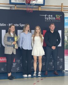 Cztery osoby pozują do zdjęcia podczas uroczystości sportowej w hali sportowej. Kobieta po lewej trzyma złoty puchar i teczkę z dokumentami. W tle znajduje się ścianka promocyjna z napisem „Lublin – Miasto Inspiracji” oraz kosz do koszykówki.