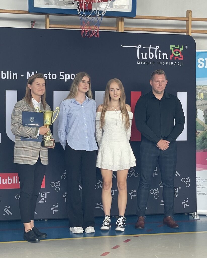 Cztery osoby pozują do zdjęcia podczas uroczystości sportowej w hali sportowej. Kobieta po lewej trzyma złoty puchar i teczkę z dokumentami. W tle znajduje się ścianka promocyjna z napisem „Lublin – Miasto Inspiracji” oraz kosz do koszykówki.
