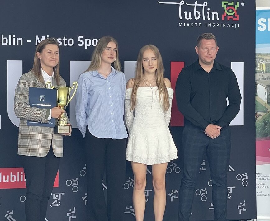 Cztery osoby pozują do zdjęcia podczas uroczystości sportowej w hali sportowej. Kobieta po lewej trzyma złoty puchar i teczkę z dokumentami. W tle znajduje się ścianka promocyjna z napisem „Lublin – Miasto Inspiracji” oraz kosz do koszykówki.