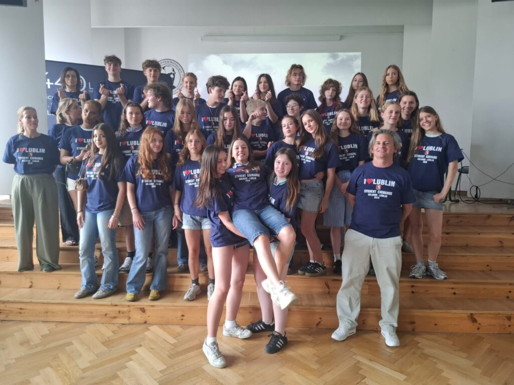 Duża grupa młodzieży i opiekunów pozuje do wspólnego zdjęcia w szkolnej sali. Uczestnicy mają granatowe koszulki z napisem „I ♥ LUBLIN – Student Exchange”. Część osób stoi na podwyższeniu, a kilka przyjmuje swobodne, żartobliwe pozy.