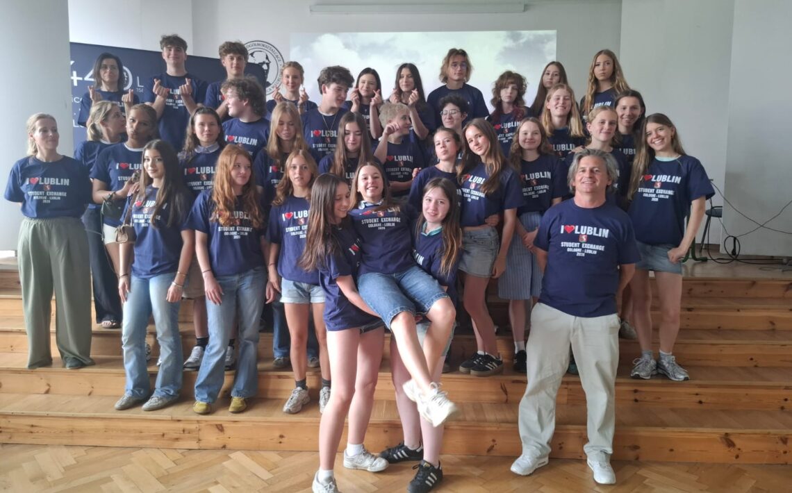 Duża grupa młodzieży i opiekunów pozuje do wspólnego zdjęcia w szkolnej sali. Uczestnicy mają granatowe koszulki z napisem „I ♥ LUBLIN – Student Exchange”. Część osób stoi na podwyższeniu, a kilka przyjmuje swobodne, żartobliwe pozy.