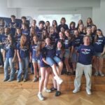 Duża grupa młodzieży i opiekunów pozuje do wspólnego zdjęcia w szkolnej sali. Uczestnicy mają granatowe koszulki z napisem „I ♥ LUBLIN – Student Exchange”. Część osób stoi na podwyższeniu, a kilka przyjmuje swobodne, żartobliwe pozy.