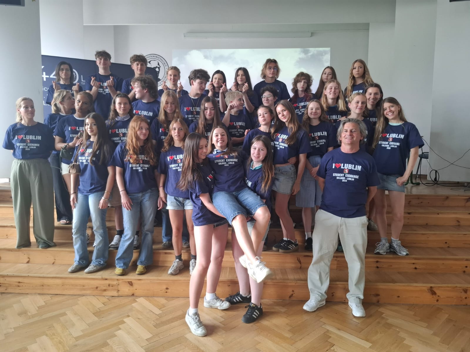 Duża grupa młodzieży i opiekunów pozuje do wspólnego zdjęcia w szkolnej sali. Uczestnicy mają granatowe koszulki z napisem „I ♥ LUBLIN – Student Exchange”. Część osób stoi na podwyższeniu, a kilka przyjmuje swobodne, żartobliwe pozy.