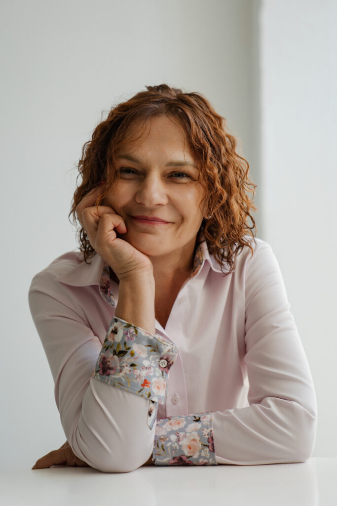 Portret uśmiechniętej kobiety o kręconych, rudych włosach, ubranej w jasnoróżową koszulę z kwiatowymi mankietami. Kobieta opiera głowę na dłoni i patrzy w stronę aparatu. Tło jest jasne, jednolite i minimalistyczne.