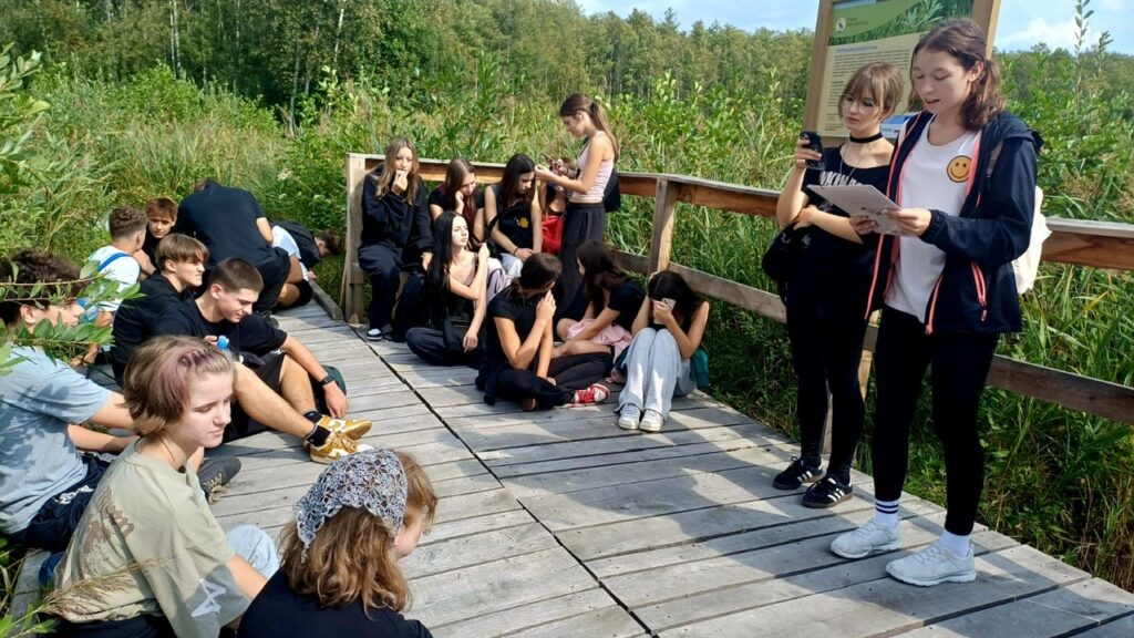 Uczniowie odpoczywają na drewnianej platformie pośród roślinności. Dwie uczennice stoją obok tablicy edukacyjnej, jedna z nich czyta informacje z kartki.