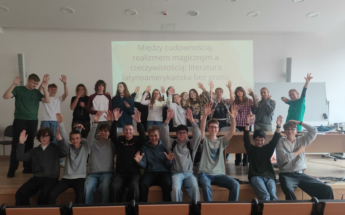 Grupowe zdjęcie młodzieży po zajęciach poświęconych literaturze latynoamerykańskiej. Uczestnicy stoją i siedzą przed ekranem prezentacji, radośnie unosząc ręce.