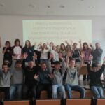 Grupowe zdjęcie młodzieży po zajęciach poświęconych literaturze latynoamerykańskiej. Uczestnicy stoją i siedzą przed ekranem prezentacji, radośnie unosząc ręce.
