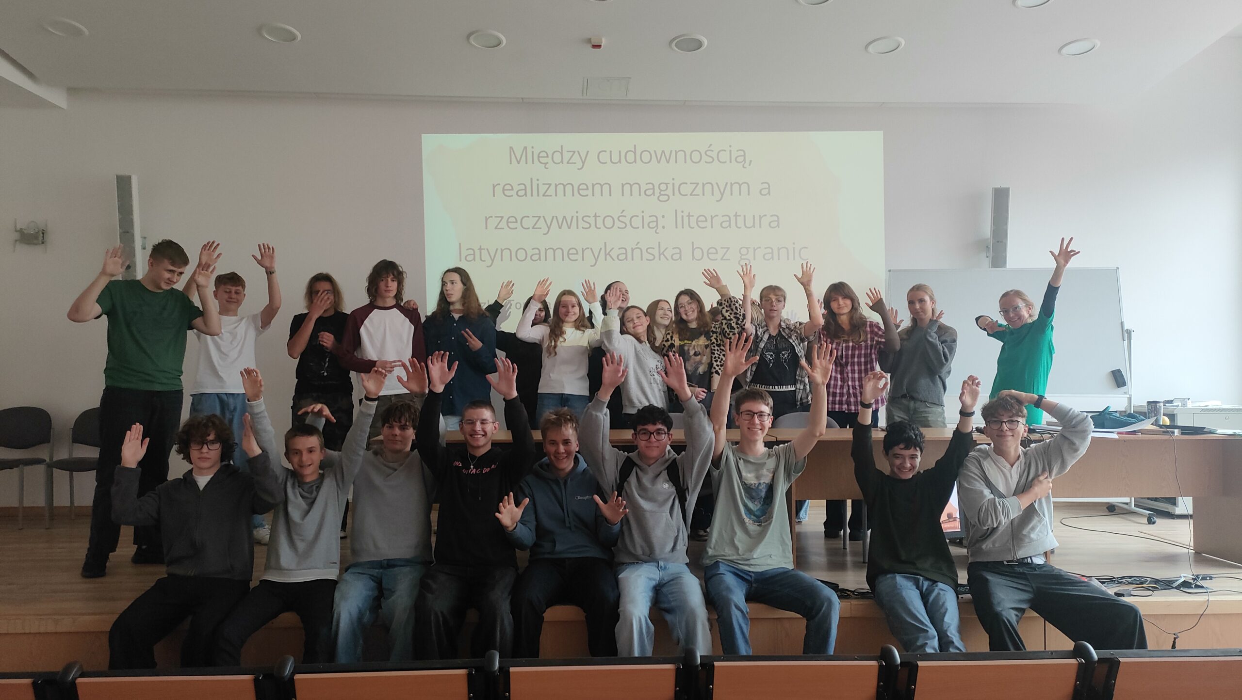 Grupowe zdjęcie młodzieży po zajęciach poświęconych literaturze latynoamerykańskiej. Uczestnicy stoją i siedzą przed ekranem prezentacji, radośnie unosząc ręce.