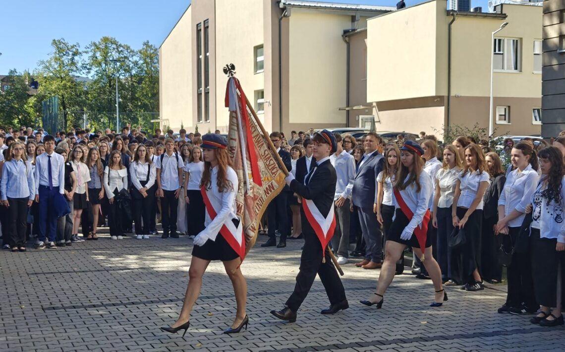 Poczet sztandarowy maszeruje przez szkolny dziedziniec przed licznie zgromadzonymi uczniami. Trzy osoby mają biało-czerwone szarfy i białe rękawiczki; środkowa niesie sztandar szkoły.