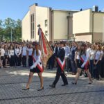 Poczet sztandarowy maszeruje przez szkolny dziedziniec przed licznie zgromadzonymi uczniami. Trzy osoby mają biało-czerwone szarfy i białe rękawiczki; środkowa niesie sztandar szkoły.