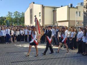 Poczet sztandarowy maszeruje przez szkolny dziedziniec przed licznie zgromadzonymi uczniami. Trzy osoby mają biało-czerwone szarfy i białe rękawiczki; środkowa niesie sztandar szkoły.