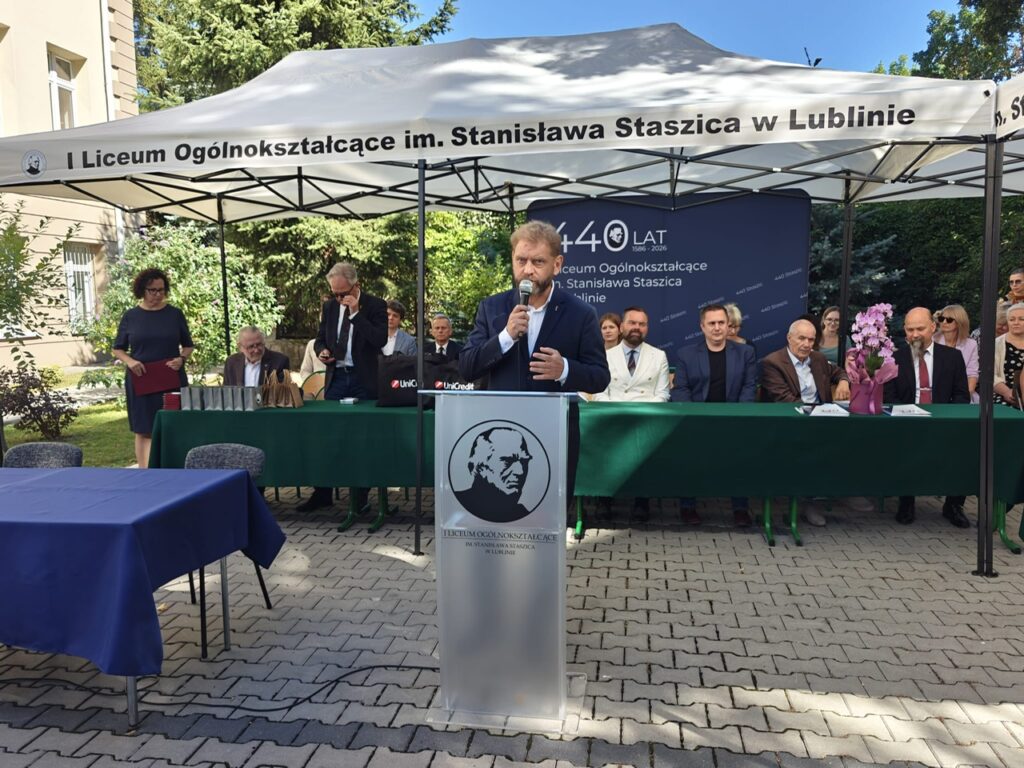 Mężczyzna w granatowym garniturze przemawia przy mównicy z wizerunkiem Stanisława Staszica. Za nim pod białym namiotem siedzą uczestnicy uroczystości przy długim stole przykrytym zielonym obrusem.