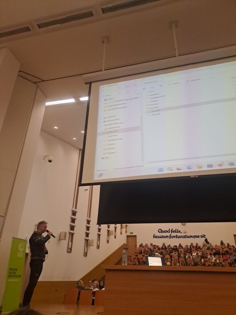 Duża aula wykładowa podczas spotkania. Po lewej stronie mężczyzna w czarnym stroju przemawia do mikrofonu. Nad nim znajduje się duży ekran projekcyjny z widokiem folderów komputera. W tle siedzi liczna publiczność.