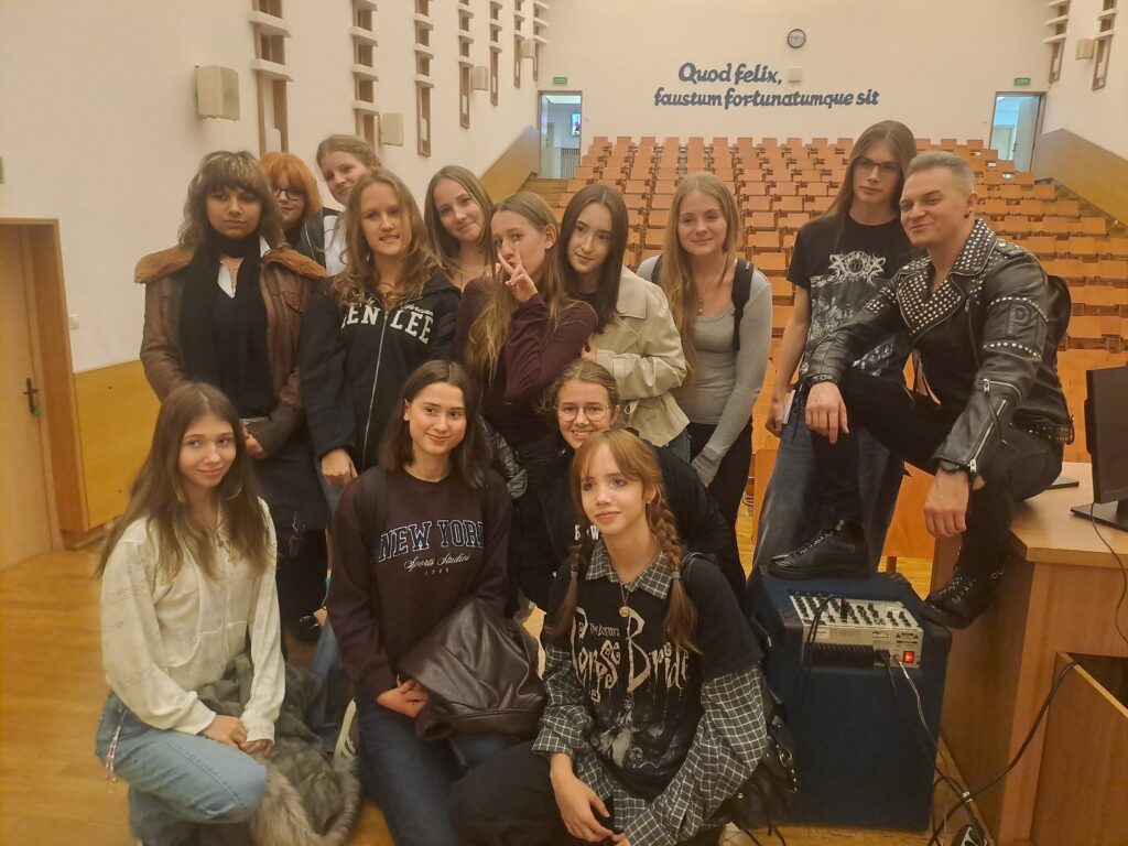 Grupowe zdjęcie w auli. Kilkanaście młodych osób pozuje razem z mężczyzną ubranym w czarną, ćwiekowaną kurtkę. W tle widać rzędy drewnianych siedzeń oraz łaciński napis na ścianie: „Quod felix, faustum fortunatumque sit”.