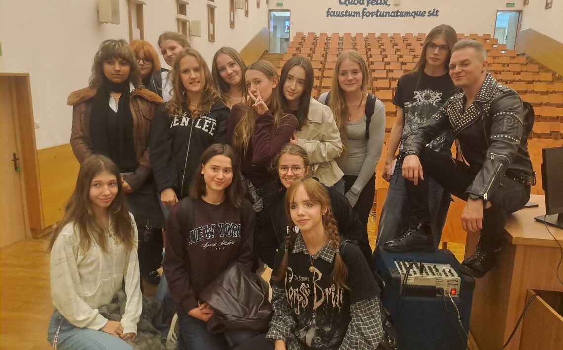 Grupowe zdjęcie w auli. Kilkanaście młodych osób pozuje razem z mężczyzną ubranym w czarną, ćwiekowaną kurtkę. W tle widać rzędy drewnianych siedzeń oraz łaciński napis na ścianie: „Quod felix, faustum fortunatumque sit”.