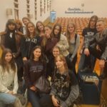 Grupowe zdjęcie w auli. Kilkanaście młodych osób pozuje razem z mężczyzną ubranym w czarną, ćwiekowaną kurtkę. W tle widać rzędy drewnianych siedzeń oraz łaciński napis na ścianie: „Quod felix, faustum fortunatumque sit”.