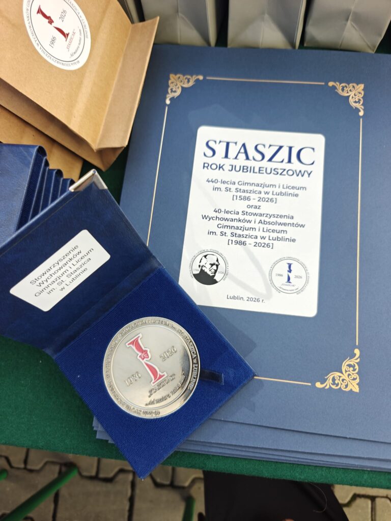 Zbliżenie jubileuszowych materiałów: granatowej publikacji „STASZIC – ROK JUBILEUSZOWY”, medalu w granatowym etui oraz papierowych toreb. Na publikacji podano jubileusz 440-lecia szkoły, lata 1586–2026, oraz 40-lecia Stowarzyszenia Wychowanków i Absolwentów, 1986–2026.