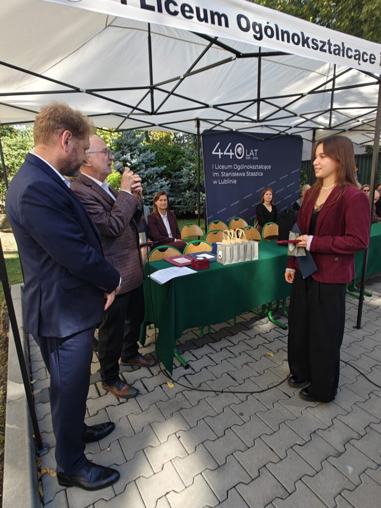 Starszy mężczyzna przemawia przez mikrofon do młodej kobiety w bordowej marynarce, stojącej naprzeciwko niego z dokumentami i pamiątkowym etui. W tle plansza jubileuszu 440-lecia szkoły.