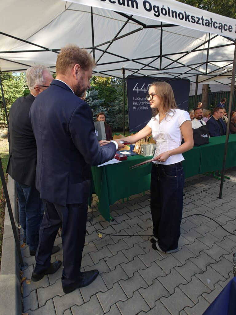 Młoda kobieta w białej bluzce i okularach odbiera dokument lub pamiątkę od mężczyzny w granatowym garniturze. W tle uczestnicy uroczystości oraz stół przykryty zielonym obrusem.