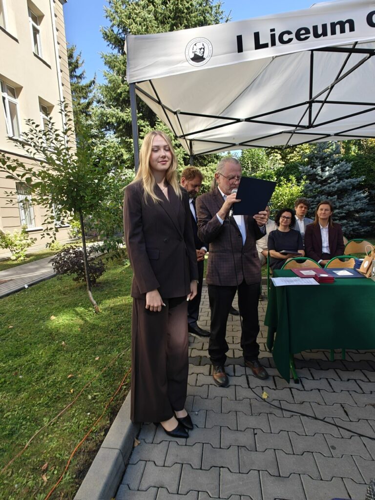 Młoda kobieta w ciemnym garniturze stoi obok starszego mężczyzny odczytującego tekst z granatowej teczki do mikrofonu. Scena odbywa się w szkolnym ogrodzie pod namiotem jubileuszowym.