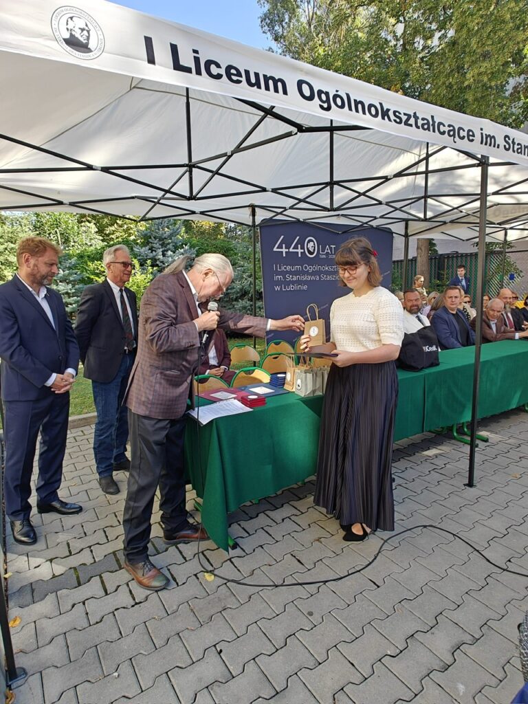 Podczas jubileuszu szkoły starszy mężczyzna z mikrofonem wręcza młodej kobiecie pamiątkowy medal w etui. Za nimi znajduje się zielony stół z dokumentami i upominkami oraz jubileuszowa plansza „440 lat”.
