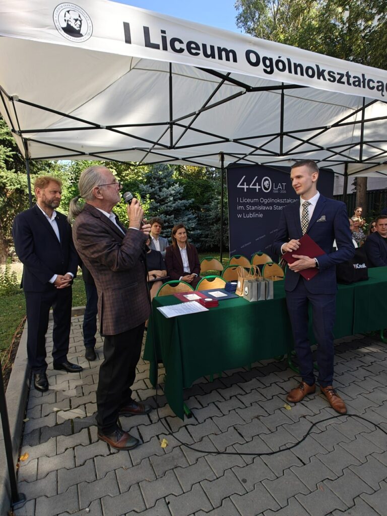 Uroczystość jubileuszowa I Liceum Ogólnokształcącego im. Stanisława Staszica w Lublinie. Pod białym namiotem starszy mężczyzna przemawia przez mikrofon do młodego mężczyzny w garniturze trzymającego bordową teczkę. W tle stół z pamiątkami i plansza „440 lat, 1586–2026”.