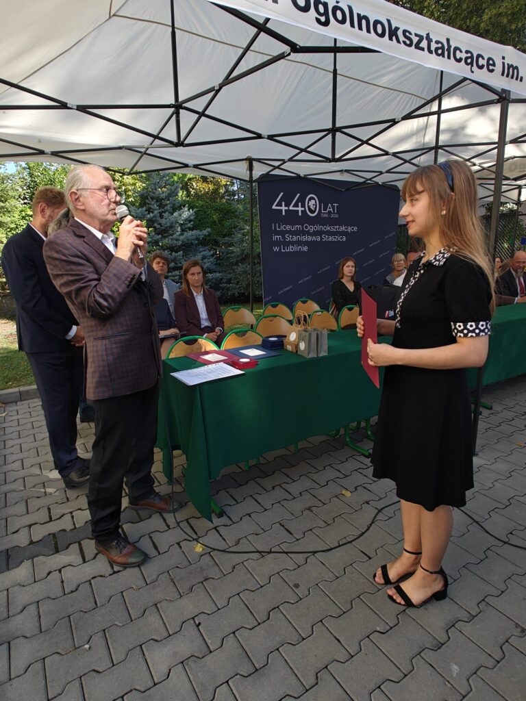 Starszy mężczyzna przemawia przez mikrofon do młodej kobiety w czarnej sukience. Kobieta stoi naprzeciwko niego, trzymając przed sobą bordową teczkę; w tle znajduje się jubileuszowy baner I Liceum Ogólnokształcącego im. Stanisława Staszica w Lublinie.