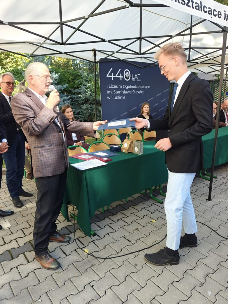 Starszy mężczyzna z mikrofonem przekazuje granatową teczkę młodemu mężczyźnie w czarnej marynarce, jasnych spodniach i okularach. Za nimi przygotowane są kolejne dyplomy i torby.