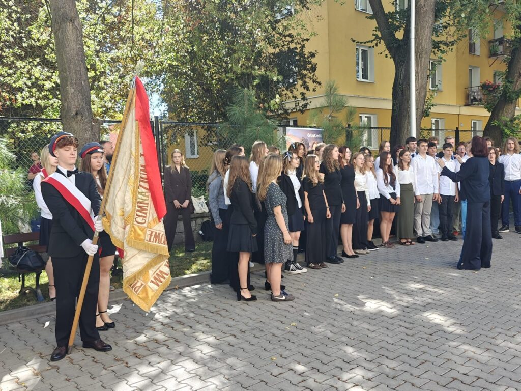 Szkolny chór ustawiony w kilku rzędach na dziedzińcu wykonuje utwór pod kierunkiem dyrygentki. Po lewej stoi poczet sztandarowy z dużym szkolnym sztandarem.
