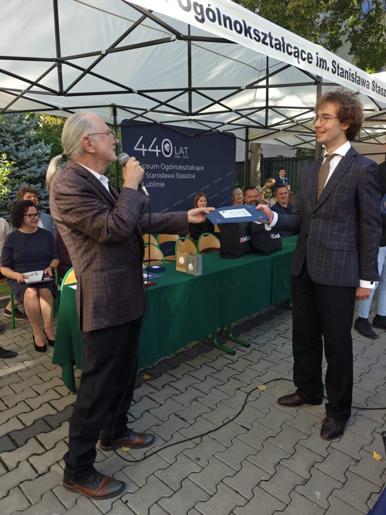 Starszy mężczyzna z mikrofonem podaje granatową teczkę młodemu mężczyźnie w ciemnym garniturze. Za nimi widoczny jest napis „440 lat” oraz nazwa szkoły.