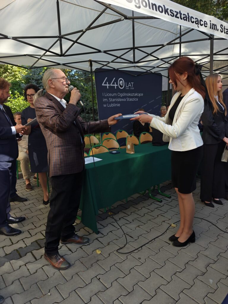 Starszy mężczyzna z mikrofonem przekazuje dokument młodej kobiecie w białej marynarce i czarnej spódnicy. W tle znajduje się zielony stół i jubileuszowy baner szkoły.