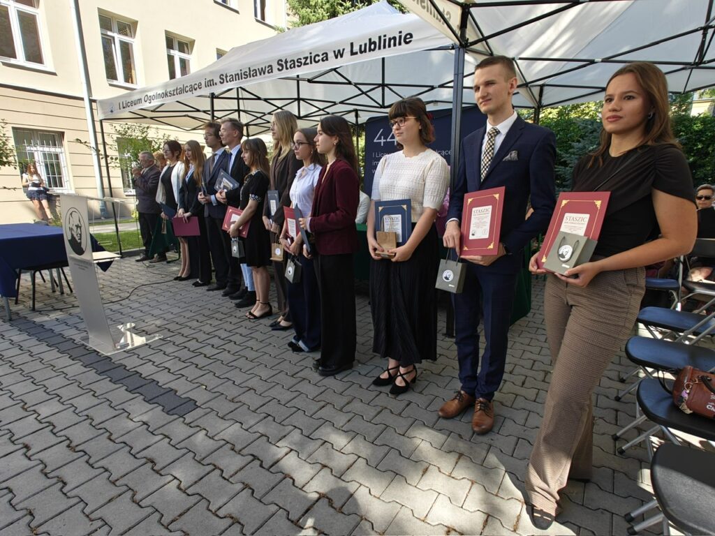 Grupa młodych osób oraz dorosłych stoi w szeregu pod szkolnym namiotem. Trzymają dyplomy, pamiątkowe plakiety, teczki i niewielkie torby prezentowe.