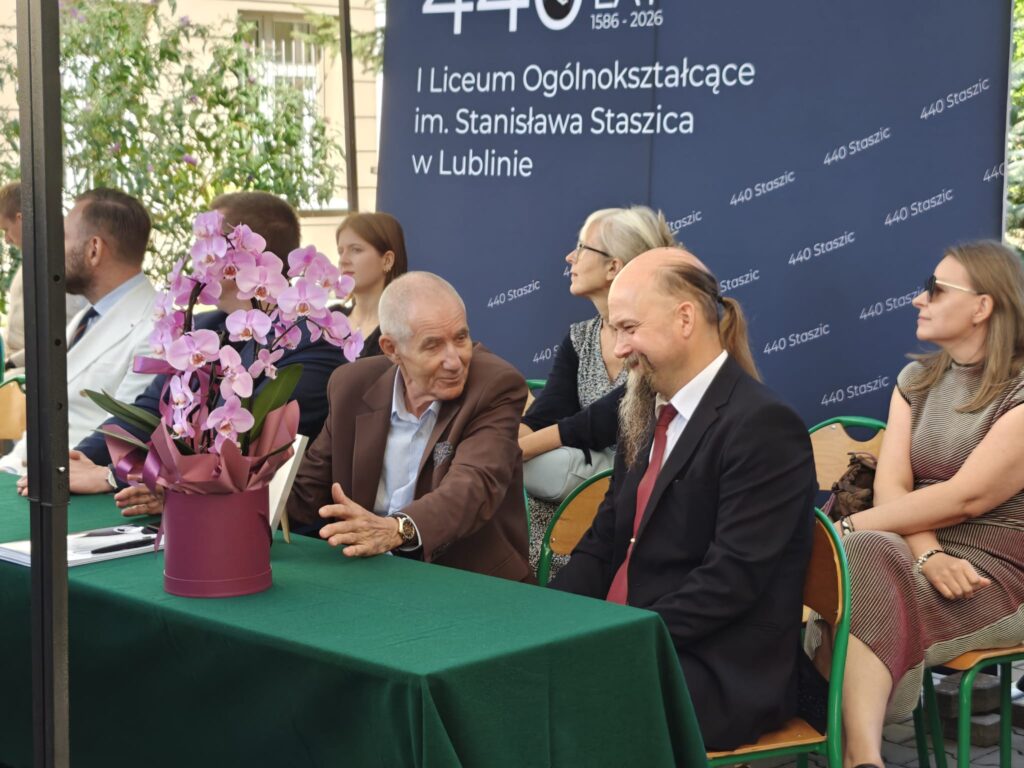 Starszy mężczyzna w brązowej marynarce rozmawia z mężczyzną w ciemnym garniturze i czerwonym krawacie. Na pierwszym planie stoi duża doniczka z różowymi storczykami, a w tle widnieje jubileuszowy baner szkoły.